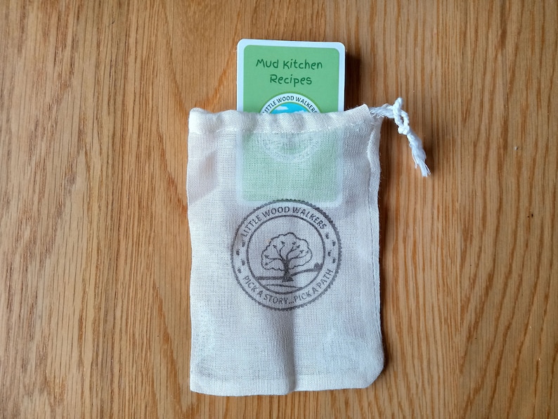 May include: A white drawstring bag with a green card inside. The card has the text "Mud Kitchen Recipes" and the logo "Little Wood Walkers Pick a Story... Pick a Path".
