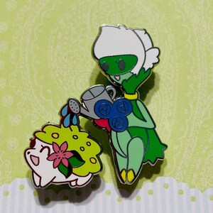 May include: A green and white cartoon character with a watering can is watering a small, white, cartoon character with a green and yellow shell. The characters are enamel pins.