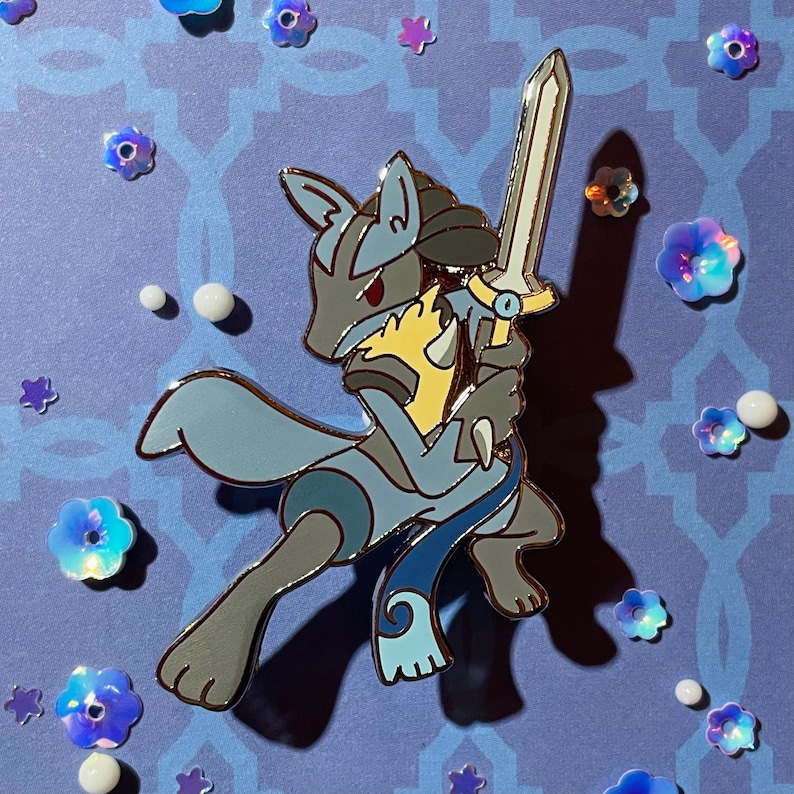 Peut inclure: Une &eacute;pingle en &eacute;mail bleue et grise repr&eacute;sentant un personnage de Pokemon tenant une &eacute;p&eacute;e. Le personnage porte une tenue bleue et jaune et une &eacute;charpe bleue et blanche.