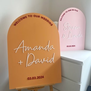 May include: Two arched welcome signs for a wedding. The sign on the left is orange with white text that reads "Welcome to our wedding, Amanda + David, 03.03.2024". The sign on the right is pink with white text that reads "Welcome to our hu plig, Steven & Linda, 03.03.2024".