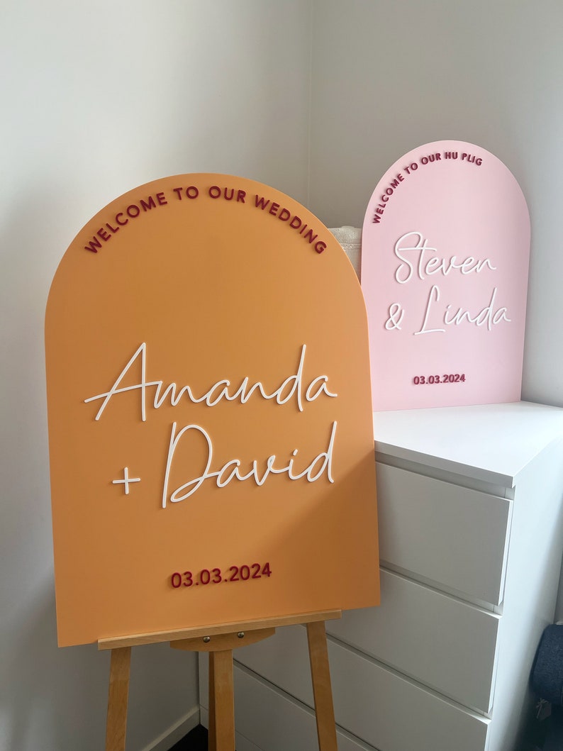 May include: Two arched welcome signs for a wedding. The sign on the left is orange with white text that reads "Welcome to our wedding, Amanda + David, 03.03.2024". The sign on the right is pink with white text that reads "Welcome to our hu plig, Steven & Linda, 03.03.2024".