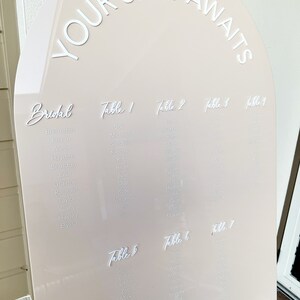 Op de afbeelding: Een beige boogvormig zitplan met de tekst "YOUR SEAT AWAITS" in witte letters. Het plan vermeldt namen voor een huwelijksreceptie, met tafels genummerd van 1 tot en met 7.