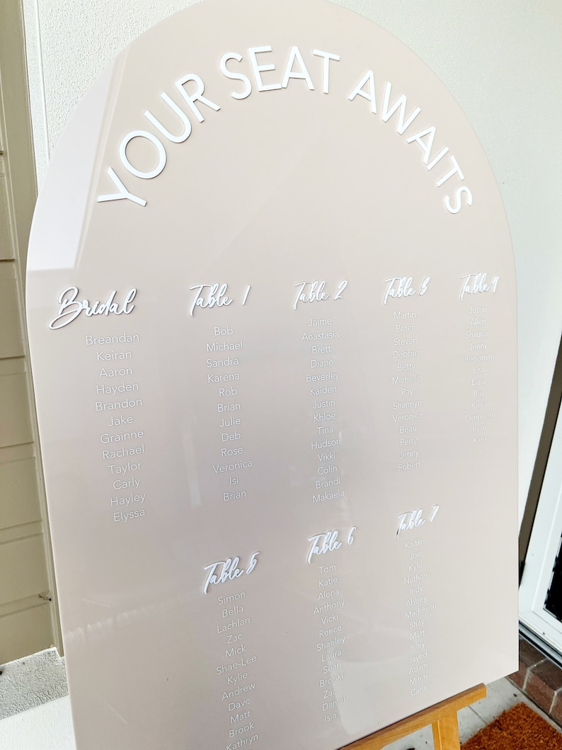 Op de afbeelding: Een beige boogvormig zitplan met de tekst "YOUR SEAT AWAITS" in witte letters. Het plan vermeldt namen voor een huwelijksreceptie, met tafels genummerd van 1 tot en met 7.