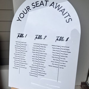 Op de afbeelding: Een wit, boogvormig bord met de tekst "YOUR SEAT AWAITS" bovenaan. Het bord vermeldt de tafels en de namen van de gasten voor een huwelijksreceptie.
