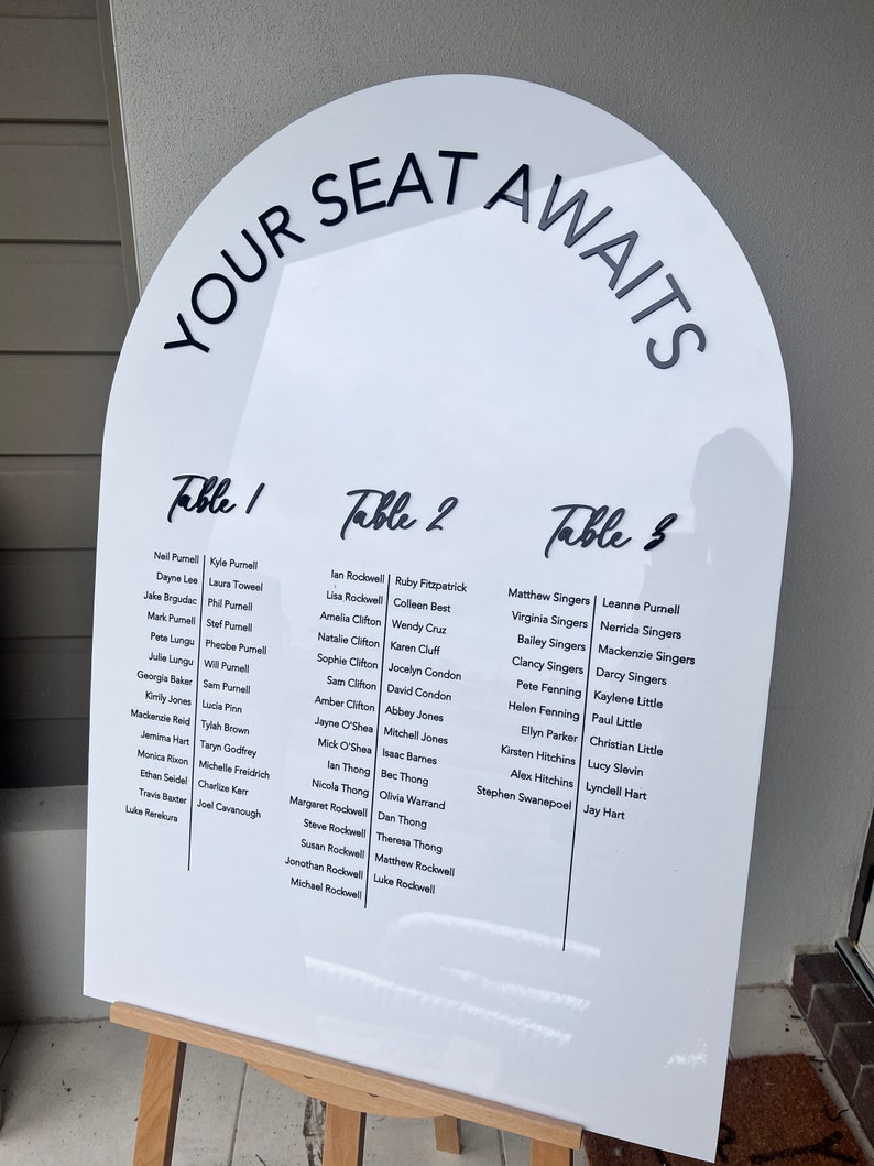 Op de afbeelding: Een wit, boogvormig bord met de tekst "YOUR SEAT AWAITS" bovenaan. Het bord vermeldt de tafels en de namen van de gasten voor een huwelijksreceptie.