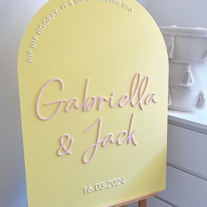 May include: A yellow arch-shaped sign with the text "Hip Hip Hooray, It's Our Wedding Day" in white lettering. The names "Gabriella & Jack" are written in pink lettering and the date "16.03.2024" is written in white lettering.