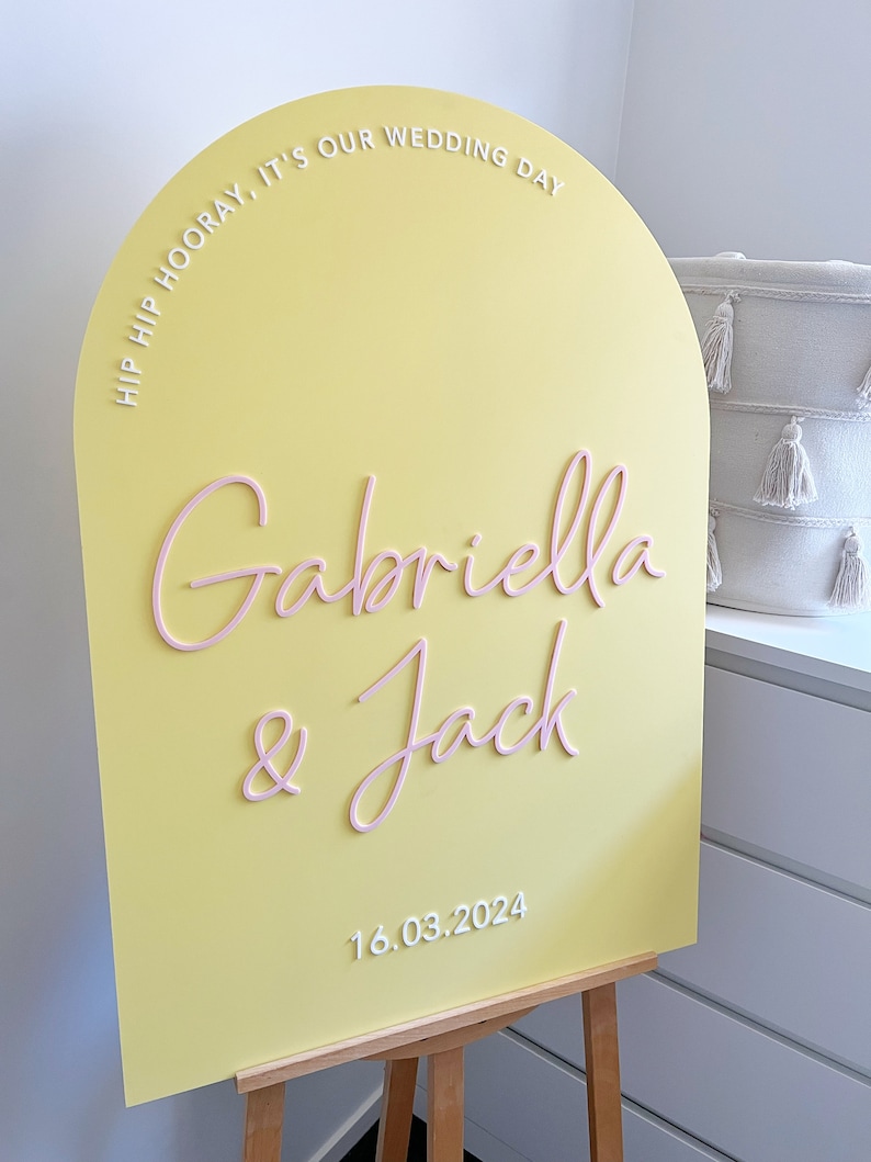 May include: A yellow arch-shaped sign with the text "Hip Hip Hooray, It's Our Wedding Day" in white lettering. The names "Gabriella & Jack" are written in pink lettering and the date "16.03.2024" is written in white lettering.