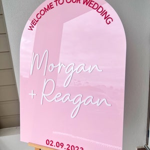 May include: Pink wedding sign with the text "Welcome to our wedding" in a red arch at the top. The names "Morgan + Reagan" are written in white cursive script. The date "02.09.2023" is written in red at the bottom.