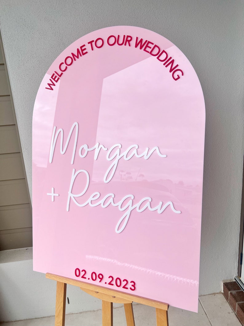 May include: Pink wedding sign with the text "Welcome to our wedding" in a red arch at the top. The names "Morgan + Reagan" are written in white cursive script. The date "02.09.2023" is written in red at the bottom.