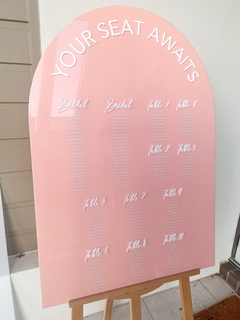 Op de afbeelding: Een roze acryl boog met de tekst "YOUR SEAT AWAITS" bovenaan. De boog is verdeeld in secties voor "Bridal" en "Table" zitplaatsverdelingen. Elke tafel heeft een lijst met namen voor de gasten.