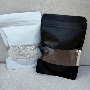 May include: Two stand-up pouches, one white and one black, each with a clear window. The white pouch contains a light-colored, textured material. The black pouch holds a darker, granular substance. Both are sealed.
