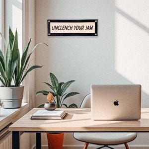 May include: A black and white sign with the text "Unclench Your Jaw" hanging on a white wall. The sign is in a home office setting with a laptop, plants, and a wooden desk.