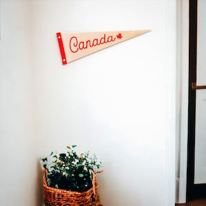 Puede incluir: Un banderín de madera con ribete rojo y una hoja de arce roja. El banderín dice "Canadá" en letras cursivas rojas.