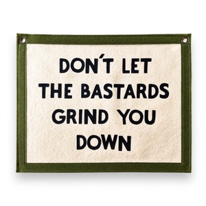 Puede incluir: Un tapiz rectangular con el texto "DON'T LET THE BASTARDS GRIND YOU DOWN" en letras negras. El fondo es blanquecino, enmarcado por un borde verde oscuro y ojales.