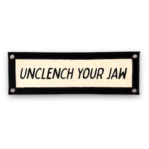 May include: Black felt banner with white fabric insert and the text "UNCLENCH YOUR JAW" in black.