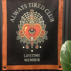 May include: Black banner with a rust-colored border and grommets. The banner features the text "ALWAYS TIRED CLUB" and "LIFETIME MEMBER" around a heart-shaped design pierced by a dagger, with a spiderweb and floral accents.