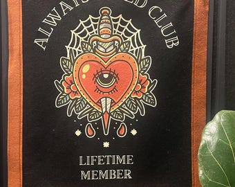May include: Black banner with a rust-coloured border and grommets. The banner features the text "ALWAYS TIRED CLUB" and "LIFETIME MEMBER" around a heart-shaped design pierced by a dagger, with a spiderweb and floral accents.