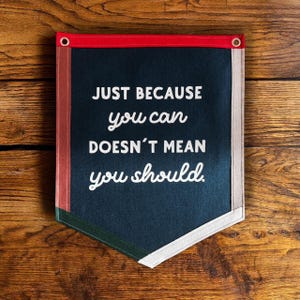 Könnte beinhalten: Ein dunkelblauer Filzbanner mit weißem Text, der "JUST BECAUSE you can DOESN'T MEAN you should." lautet. Der Banner hat einen roten, weißen und grünen Besatz und zwei Ösen oben.
