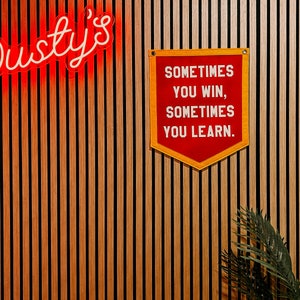 Puede incluir: Un letrero de neón rojo que dice "Dusty's" en una pared de paneles de madera. Una pancarta roja con ribete amarillo cuelga de la pared y dice "Sometimes you win, sometimes you learn."