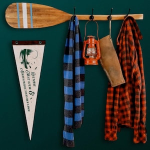 May include: A wooden oar with blue and white stripes, a pennant with text, a blue and gray striped scarf, an orange lantern, a brown leather sleeve, and a red and black plaid shirt hanging on a wooden rack with black hooks. The pennant reads "Love many, trust a few, & always paddle your own canoe."