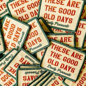 May include: A pile of embroidered patches, each featuring the phrase "THESE ARE THE GOOD OLD DAYS" in orange. The patches have a cream-colored background, a green border, and the words "Dusty Pennants" are also visible.