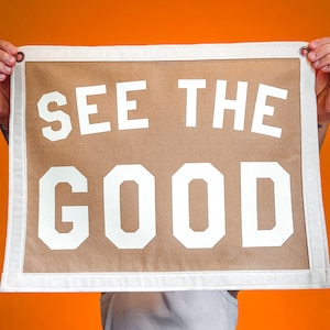Puede incluir: Una pancarta de lona marrón con texto blanco que dice "SEE THE GOOD". La pancarta está sostenida por las manos de una persona.