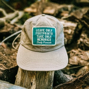 Puede incluir: Una gorra de béisbol de color beige con un parche verde que dice "Leave Only Footprints Take Only Memories Dusty Pennants".