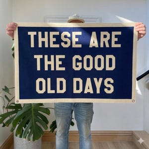 Könnte beinhalten: Ein marineblaues Banner mit dem Text "THESE ARE THE GOOD OLD DAYS" in großen, cremefarbenen Blockbuchstaben. Das Banner hat einen cremefarbenen Rand und Ösen in den Ecken. Das Banner wird von einer Person gehalten.