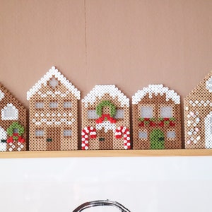 May include: Five gingerbread house ornaments made from brown and white beads. Each house has unique details, including wreaths, candy canes, and snow-covered roofs. They are arranged on a wooden shelf against a neutral background.