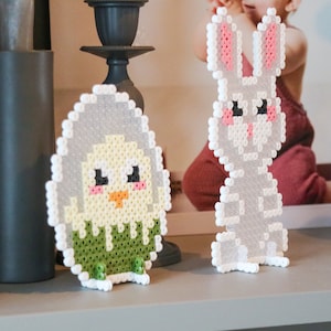 May include: Two Easter decorations made from small, colourful beads. One is a chick emerging from an egg, and the other is a bunny. The chick is yellow, green, and white, while the bunny is grey with pink accents.