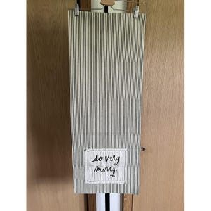 以下が含まれることがあります： 緑と白のストライプが交互に並んだ、縦縞のキッチンタオル。タオルの下部には、黒い筆記体で「so very merry」と書かれた白い長方形のパッチが付いています。