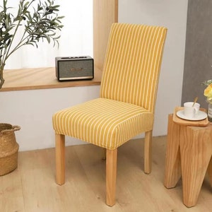 May include: A yellow and white striped dining chair. The chair has a high back and a cushioned seat. The legs are made of light-coloured wood. The chair is in a bright room, with a wooden side table.