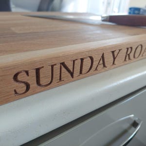 May include: A light brown wooden chopping board with the words "SUNDAY ROAST" carved into the side. A knife with a brown handle is on the board. The board is made of wood.