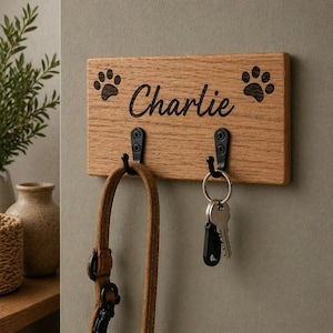 May include: A wooden key holder with the name "Charlie" in black script, paw prints, and two black hooks. A brown dog lead hangs from one hook, and keys hang from the other. The key holder is mounted on a neutral-coloured wall.