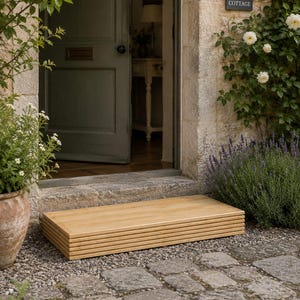 May include: A natural wood step is positioned on a stone path leading to a doorway. The step is rectangular, constructed from horizontal wooden planks. The open door reveals a glimpse of the interior. A sign on the wall reads "Rose Cottage."