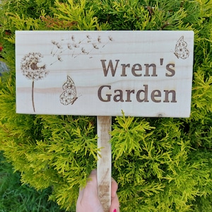 May include: A wooden garden sign with the text "Wren's Garden" burned into the surface. The sign is decorated with a dandelion and butterflies.