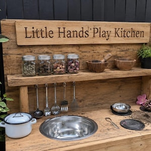 May include: A wooden play kitchen featuring a backboard with the text Little Hands Play Kitchen, a shelf holding four glass jars, a wooden bowl, and a small potted plant. The counter includes a metal sink, a white pot, and metal utensils.