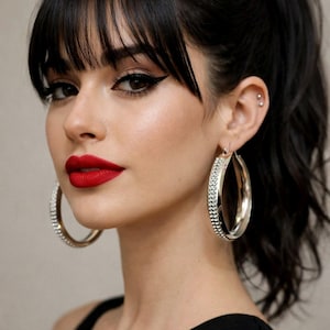 Puede incluir: Una mujer con cabello oscuro y flequillo, usando grandes pendientes de aro plateados con piedras transparentes. Tiene lápiz labial rojo y delineador de ojos alado. Su cabello está recogido en una cola de caballo.