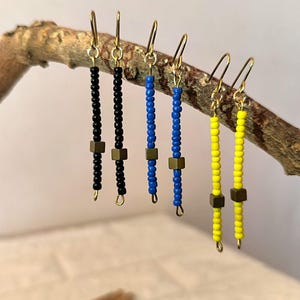 Può includere: Orecchini fatti a mano in nero, blu e giallo. Ogni orecchino presenta un gancio color oro, una fila di piccole perline e un accento metallico quadrato. Gli orecchini sono esposti su un ramo di legno naturale.
