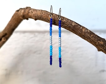 May include: Two pairs of long, beaded earrings. Each earring is a vertical strand of small beads in shades of blue, silver, and white. The earrings are hanging from a small branch against a blurred background.