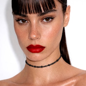 Può includere: Un ritratto in primo piano di una persona che indossa una collana girocollo di perline nere. La persona ha i capelli scuri con la frangia, occhi marroni e rossetto rosso. Lo sfondo è bianco.