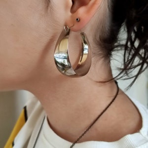 May include: Silver hoop earrings with a slightly curved shape. The earrings are large and have a shiny finish.