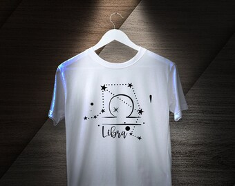 Puede incluir: Camiseta blanca con un diseño negro de constelación zodiacal de Libra. El diseño incluye un símbolo de Libra estilizado, estrellas y la palabra "Libra".