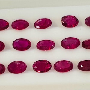 Könnte beinhalten: Eine Sammlung von fünfzehn losen, oval geschliffenen Rubinen, angeordnet in drei Reihen zu je fünf Stück. Jeder Edelstein zeigt eine tiefe, leuchtende rote Farbe mit facettierten Oberflächen, die das Licht reflektieren und eine einheitliche Form und Größe aufweisen.