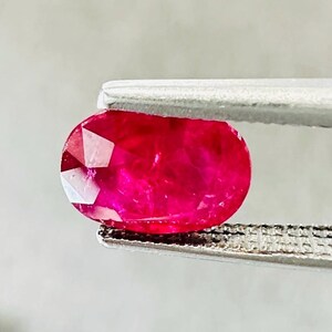 Könnte beinhalten: Ein facettierter, ovaler Rubin-Edelstein, gehalten von einer silbernen Pinzette. Der Rubin zeigt eine lebendige, tiefrote Farbe mit sichtbaren inneren Facetten, die das Licht reflektieren. Der Hintergrund ist ein weiches, neutrales Grau.