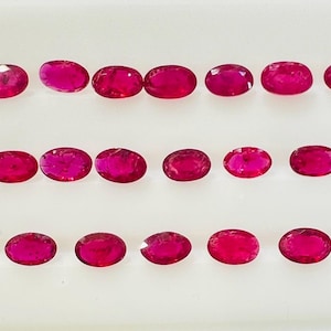 Könnte beinhalten: Eine Anordnung von oval geschliffenen Rubinen, präsentiert in ordentlichen Reihen vor einem weißen Hintergrund. Die Edelsteine weisen eine lebendige rote Farbe auf, mit unterschiedlicher Sättigung. Die Anordnung betont Form und Farbe der Edelsteine.