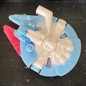 Puede incluir: Una vela con forma de nave espacial azul, blanca y roja. La vela tiene la forma del Halcón Milenario de Star Wars.