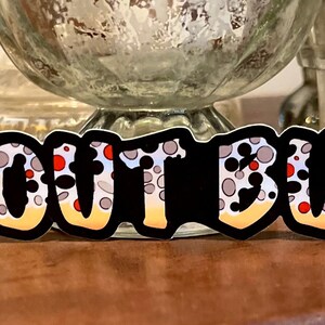 May include: A sticker with the words "TROUT BUM" in a bold, stylized font. The letters are black with a pattern of gray and red spots, and a yellow gradient. The sticker is likely for fishing enthusiasts.