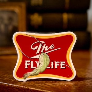 May include: A red and white sticker with the text "The Fly Life" and a green and white fly tied on a hook.