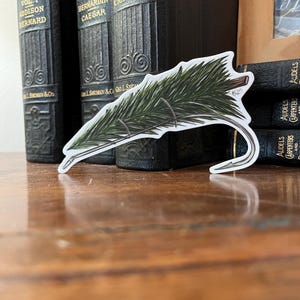 Könnte beinhalten: Ein Aufkleber mit einem grünen Kiefernbaumzweig, an dem ein Angelhaken befestigt ist. Der Aufkleber befindet sich auf einem Stapel alter Bücher.